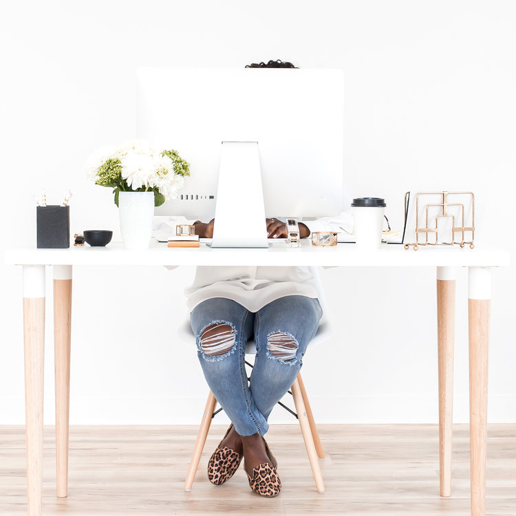 A bright white desk with flowers, a computer, coffee, and a woman seated behind it